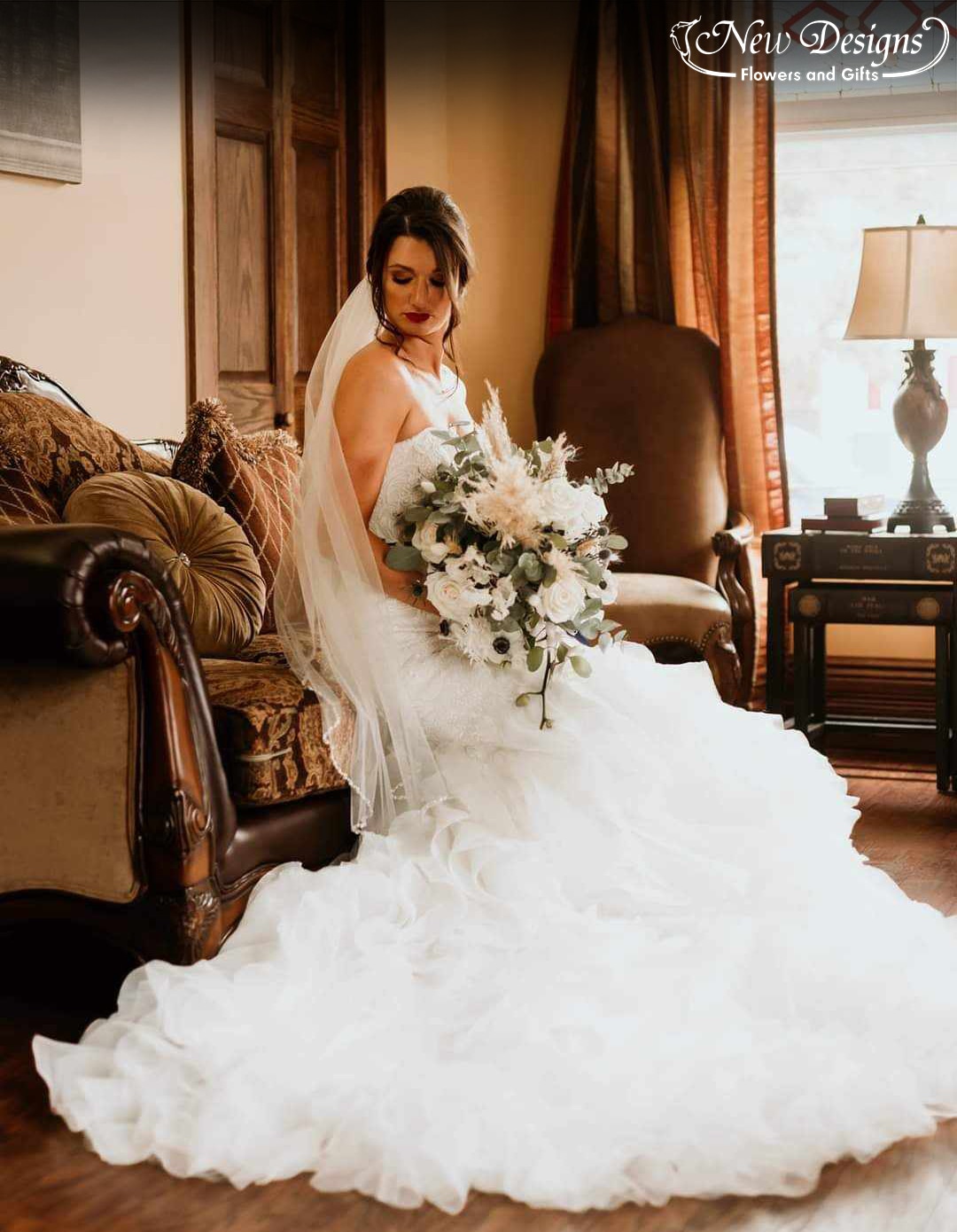 A bride sits in an antique chair in a heritage home, holding a bohemian style bouquet of wild greens, pampas grass, and white flowers including white anenomes, roses, and more